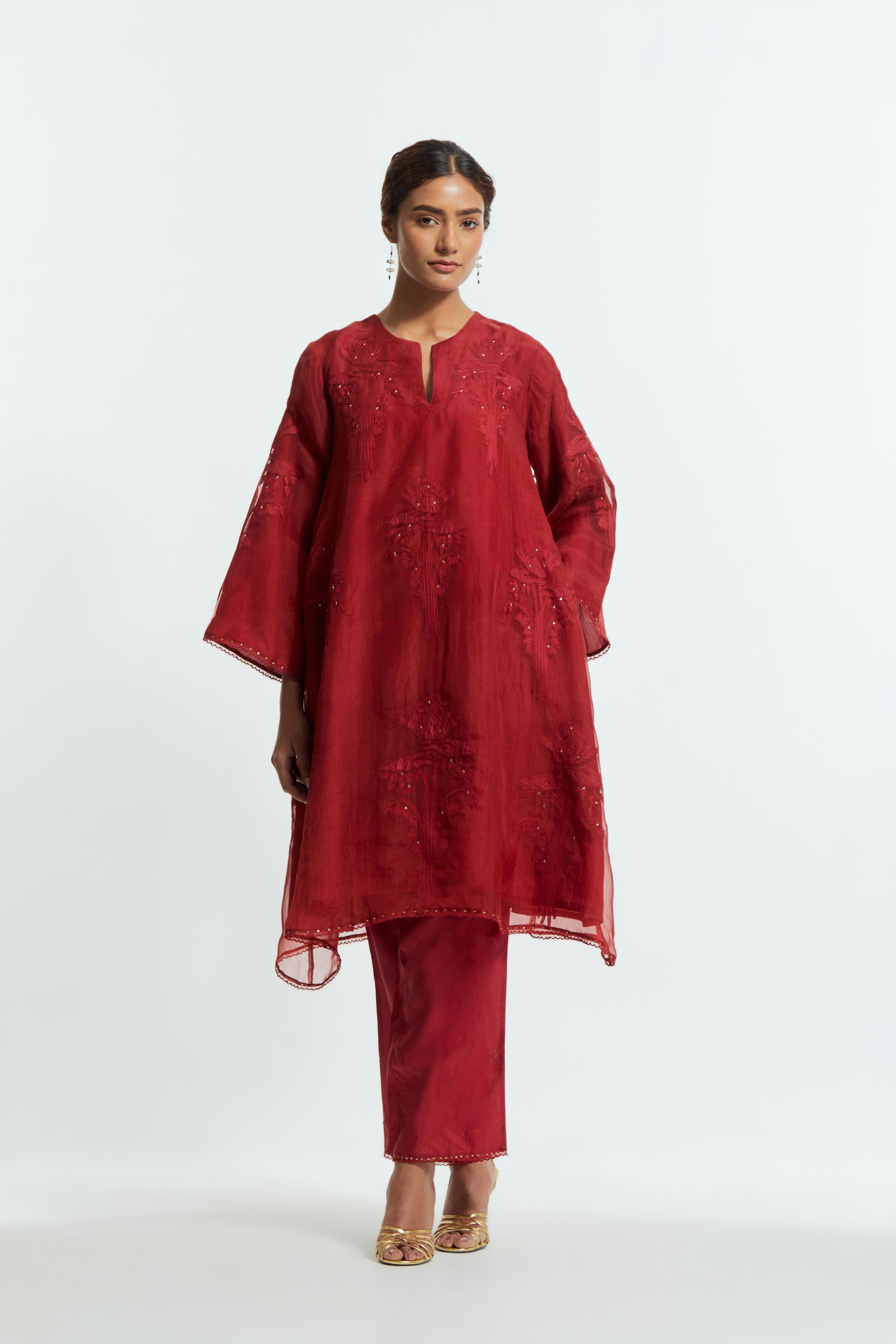 Red silk organza A-line short kurta adorned with delicately placed floral appliqué motifs and subtle sequin embellishments. Paired with red silk straight pants detailed with scalloped organza edging at the hem, highlighted with gold sequins work.