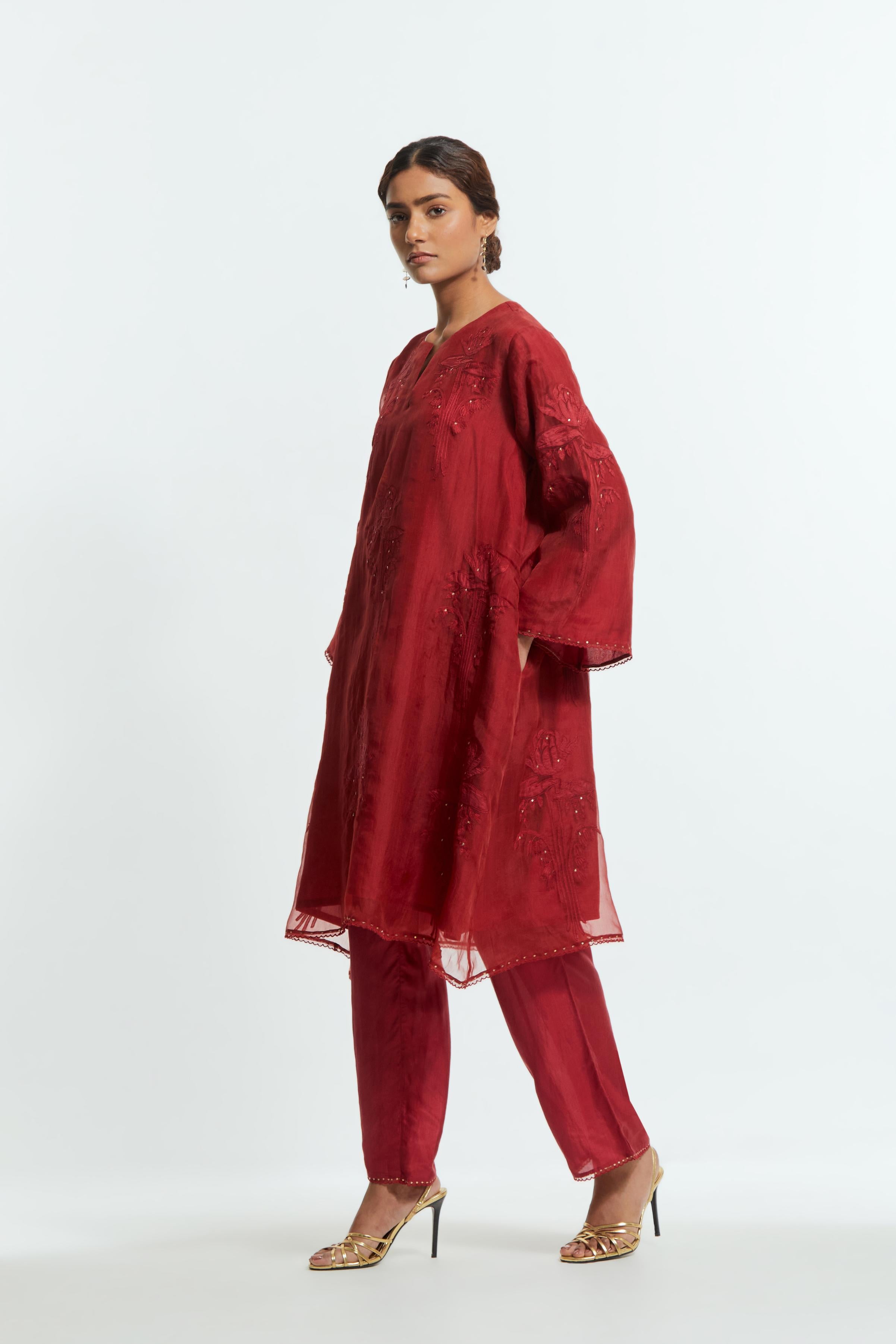 Red silk organza A-line short kurta adorned with delicately placed floral appliqué motifs and subtle sequin embellishments. Paired with red silk straight pants detailed with scalloped organza edging at the hem, highlighted with gold sequins work.