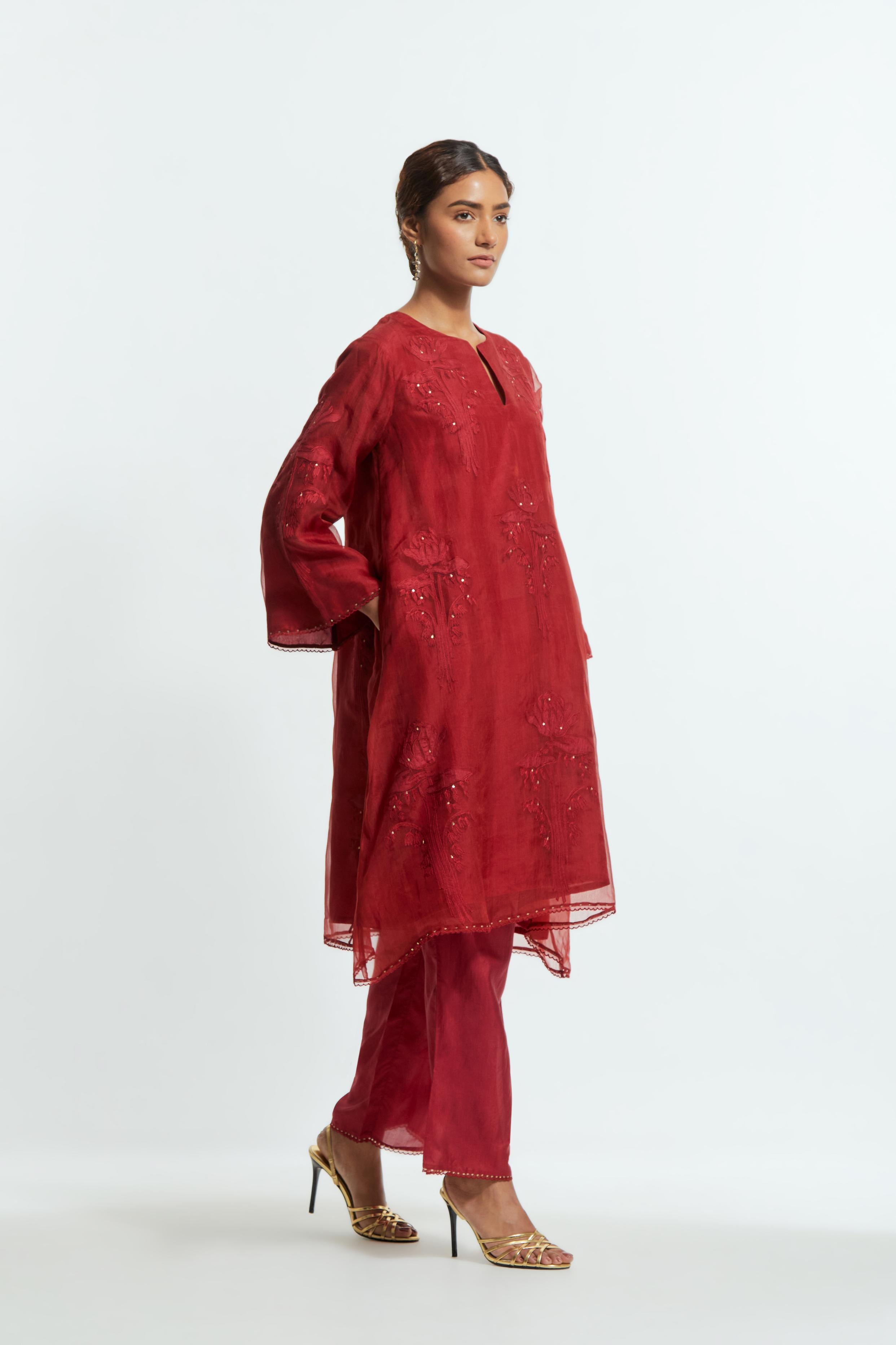Red silk organza A-line short kurta adorned with delicately placed floral appliqué motifs and subtle sequin embellishments. Paired with red silk straight pants detailed with scalloped organza edging at the hem, highlighted with gold sequins work.