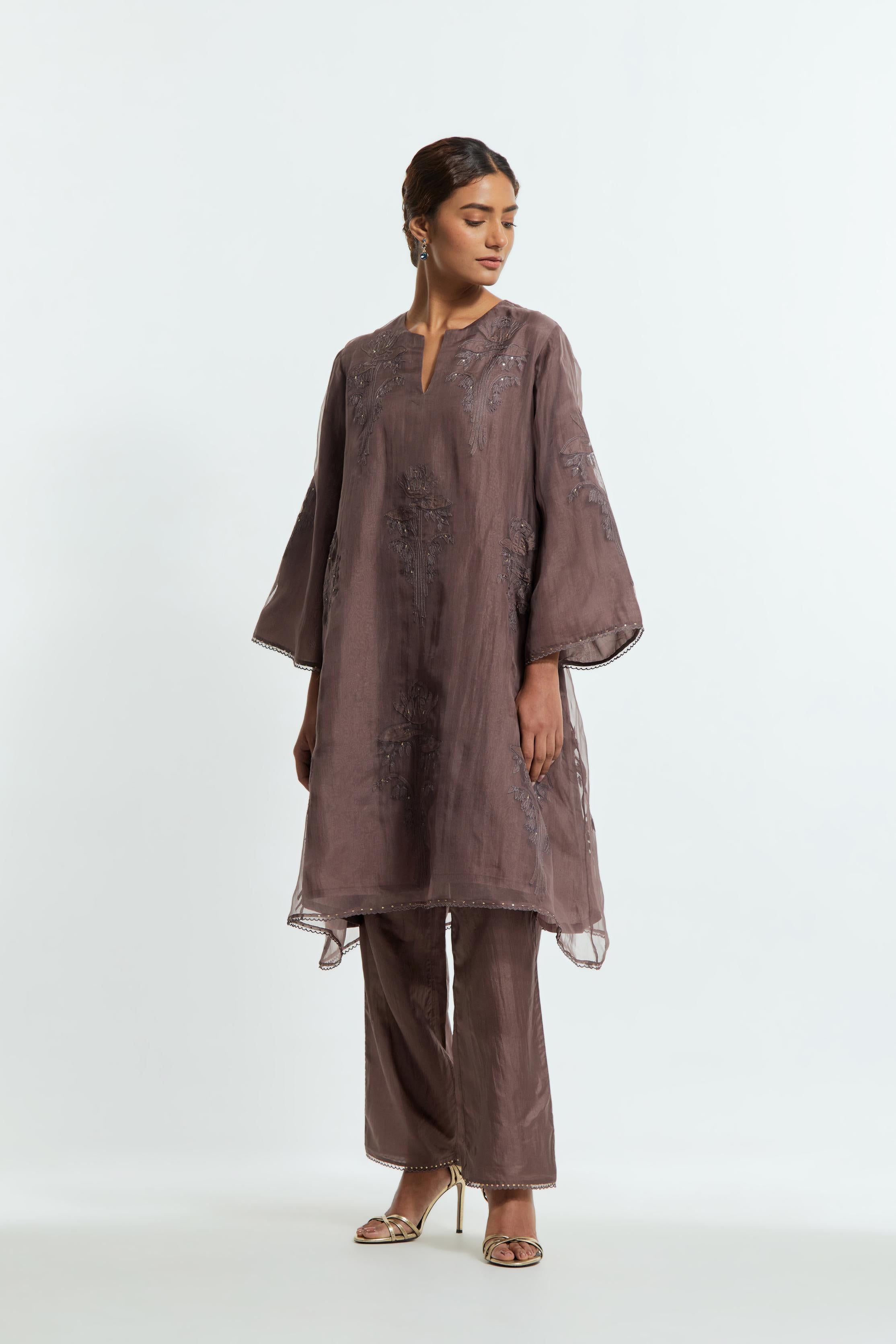 Grey silk organza A-line short kurta adorned with delicately placed floral appliqué motifs and subtle sequin embellishments. Paired with grey silk straight pants detailed with scalloped organza edging at hem, delicately highlighted with gold sequins work.