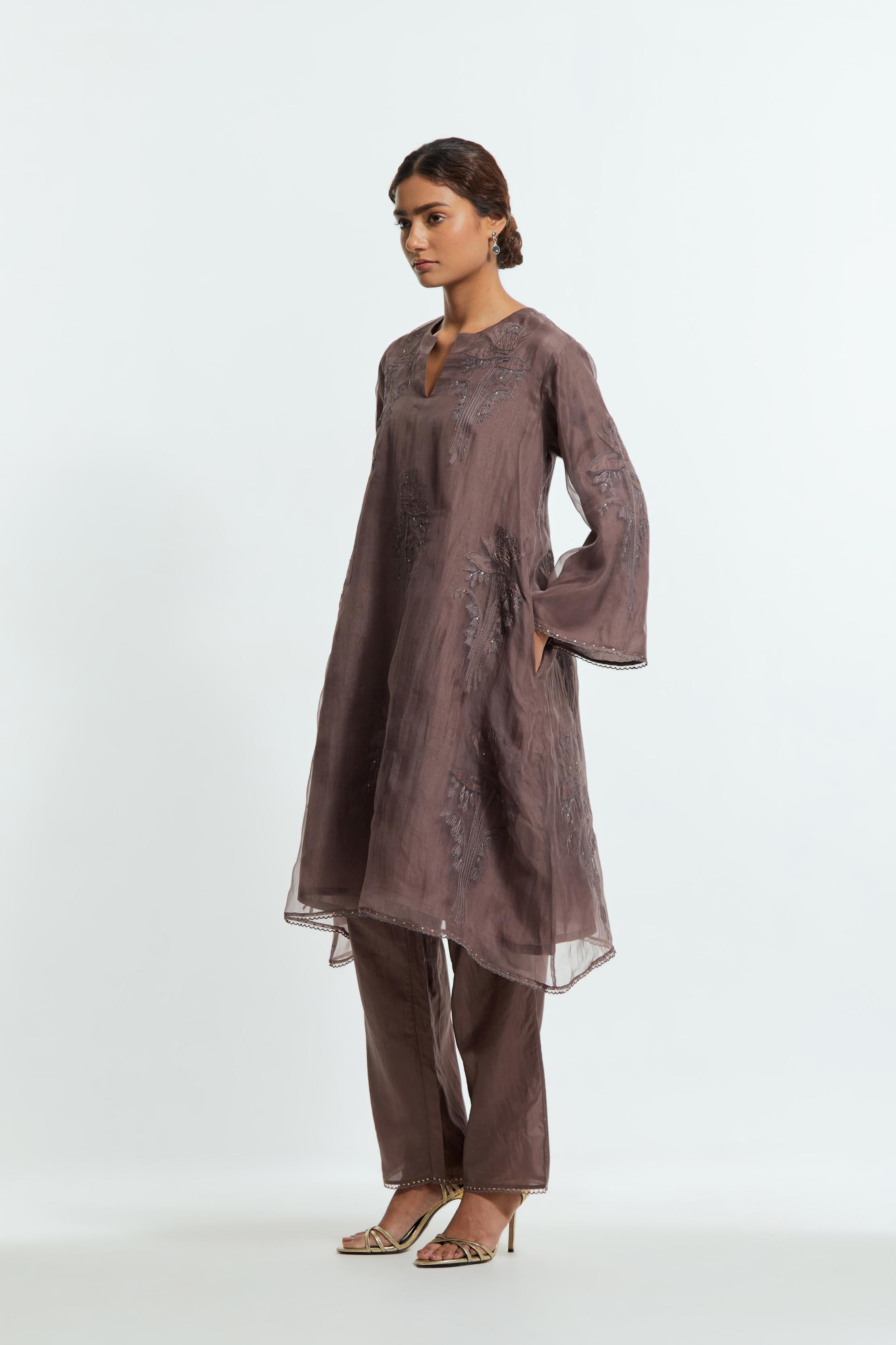 Grey silk organza A-line short kurta adorned with delicately placed floral appliqué motifs and subtle sequin embellishments. Paired with grey silk straight pants detailed with scalloped organza edging at hem, delicately highlighted with gold sequins work.