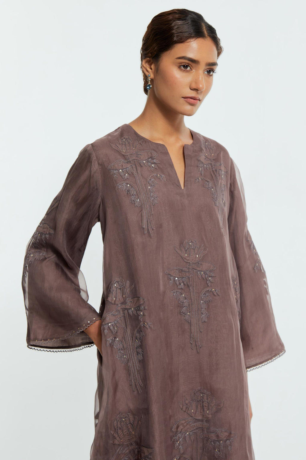 Grey silk organza A-line short kurta adorned with delicately placed floral appliqué motifs and subtle sequin embellishments. Paired with grey silk straight pants detailed with scalloped organza edging at hem, delicately highlighted with gold sequins work.