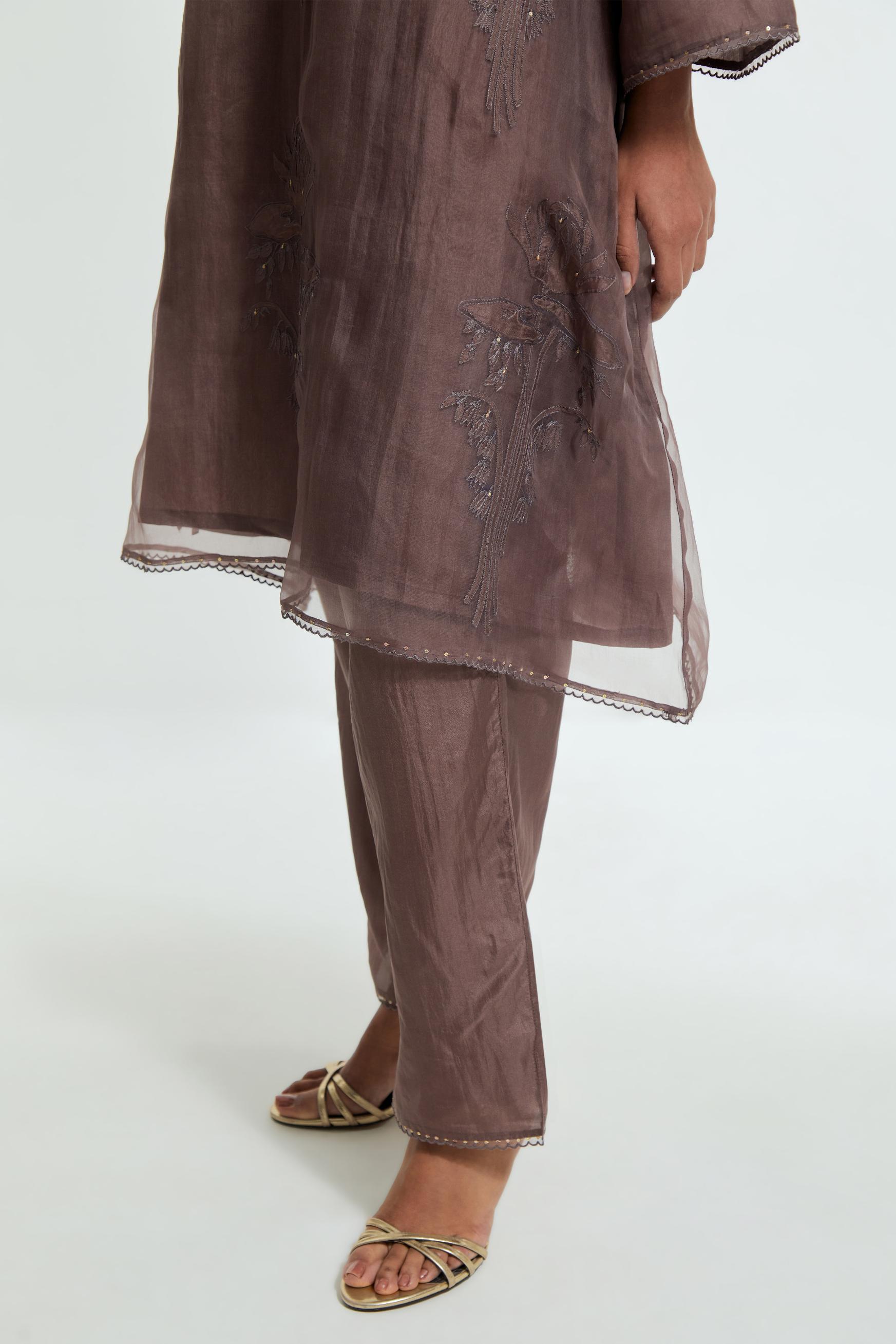 Grey silk organza A-line short kurta adorned with delicately placed floral appliqué motifs and subtle sequin embellishments. Paired with grey silk straight pants detailed with scalloped organza edging at hem, delicately highlighted with gold sequins work.