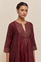 Maroon silk kurta set with asymmetric hem and hand-embroidered leaf motifs with delicate sequins work at the neck, sleeves, and hem, with sequins border running along the edges.