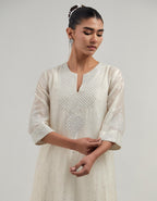Off-white silk Chanderi kurta set with asymmetric hem and hand-embroidered leaf motifs with delicate mirror and sequins work at the neck, sleeves, and hem, with a mirror border running along the edges.