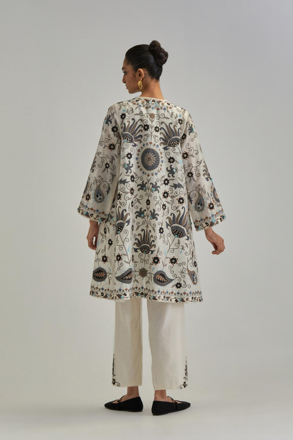 Off White short front open A-line jacket, fully embroidered with bold appliqué flowers, earthy-coloured Aari thread work and silk tassels, paired with Off white silk Chanderi straight pants with earthy coloured appliqué at hem.