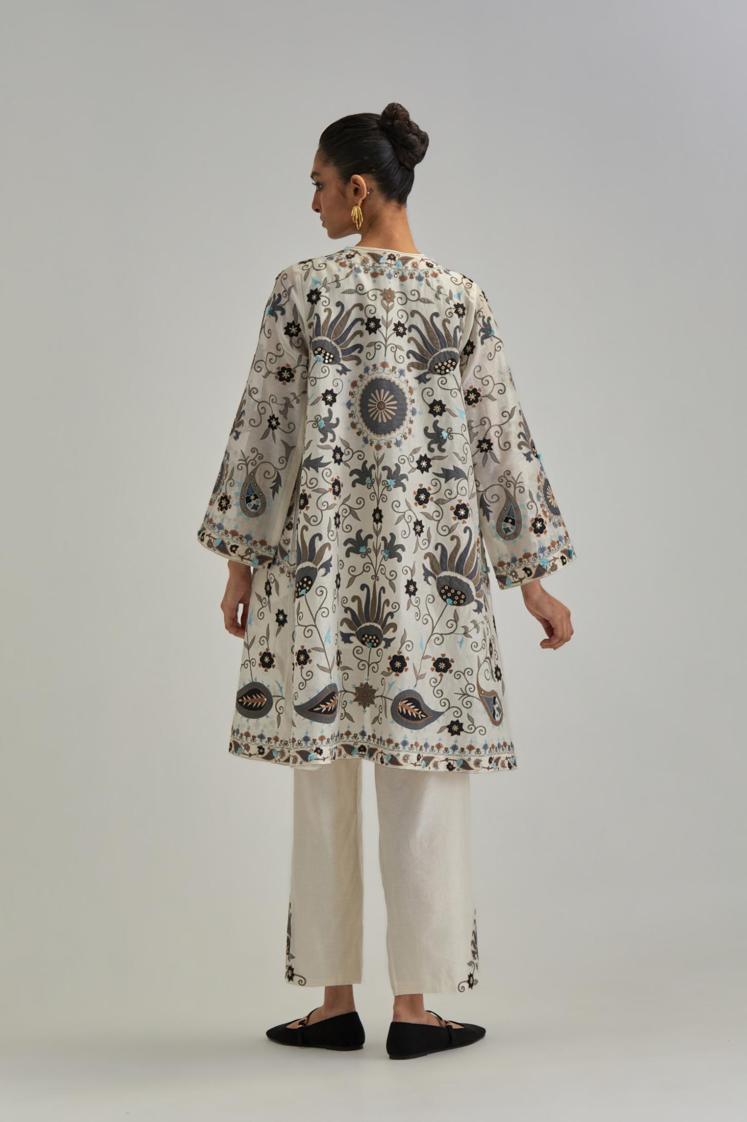 Off White short front open A-line jacket, fully embroidered with bold appliqué flowers, earthy-coloured Aari thread work and silk tassels, paired with Off white silk Chanderi straight pants with earthy coloured appliqué at hem.