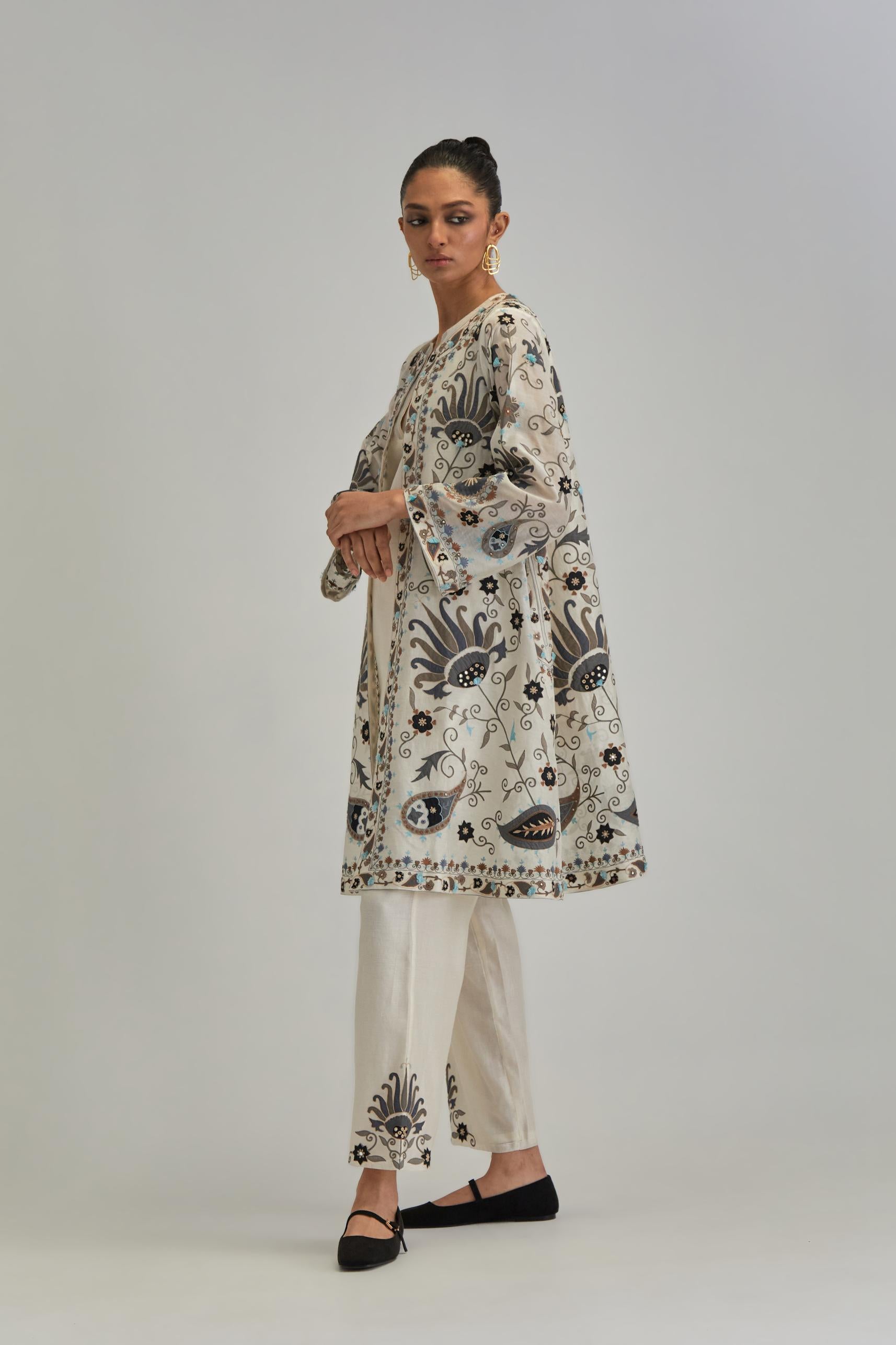 Off White short front open A-line jacket, fully embroidered with bold appliqué flowers, earthy-coloured Aari thread work and silk tassels, paired with Off white silk Chanderi straight pants with earthy coloured appliqué at hem.