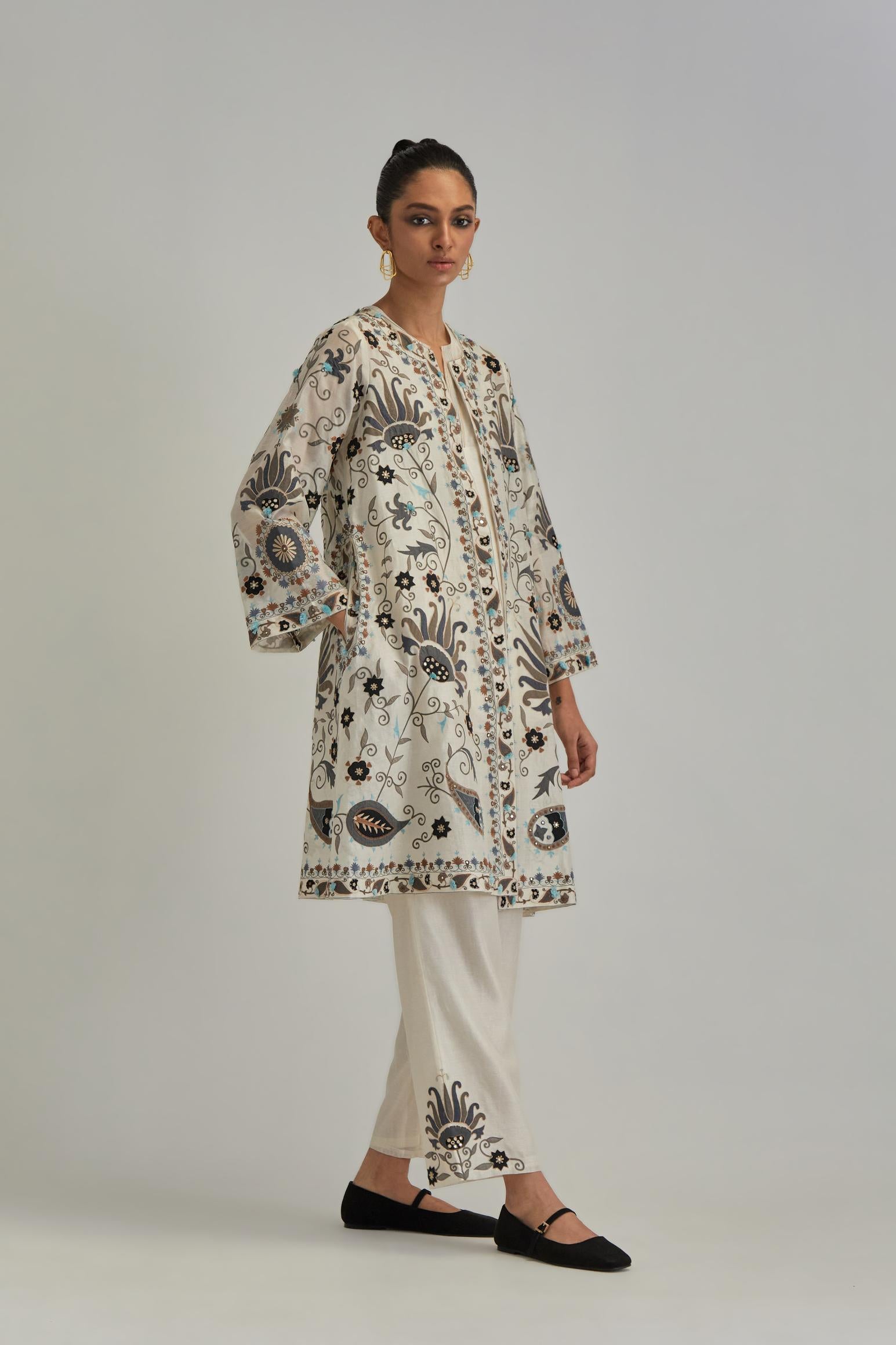 Off White short front open A-line jacket, fully embroidered with bold appliqué flowers, earthy-coloured Aari thread work and silk tassels, paired with Off white silk Chanderi straight pants with earthy coloured appliqué at hem.
