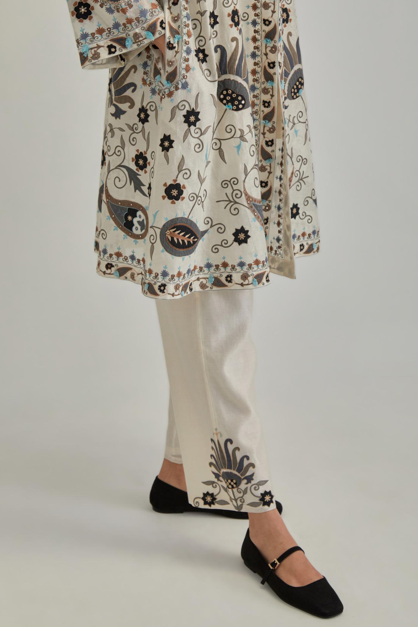 Off White short front open A-line jacket, fully embroidered with bold appliqué flowers, earthy-coloured Aari thread work and silk tassels, paired with Off white silk Chanderi straight pants with earthy coloured appliqué at hem.