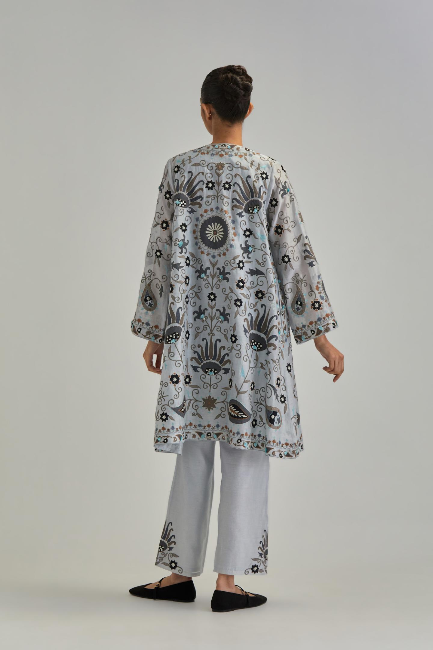 Blue short front open A-line jacket, fully embroidered with bold appliqué flowers, earthy-coloured Aari thread work and silk tassels, paired with Blue silk Chanderi straight pants with earthy coloured appliqué at hem.