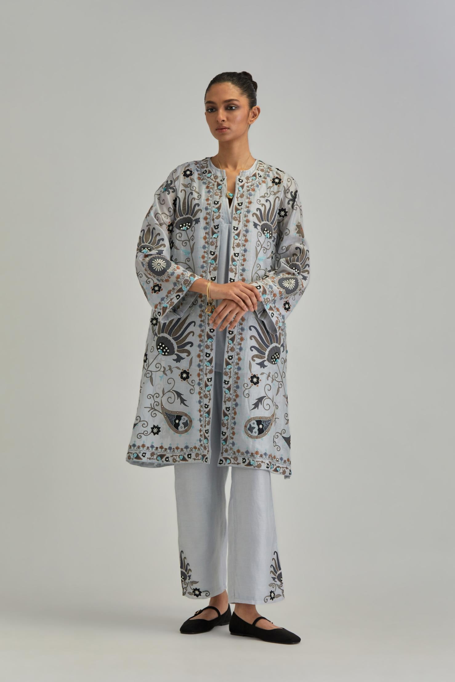 Blue short front open A-line jacket, fully embroidered with bold appliqué flowers, earthy-coloured Aari thread work and silk tassels, paired with Blue silk Chanderi straight pants with earthy coloured appliqué at hem.
