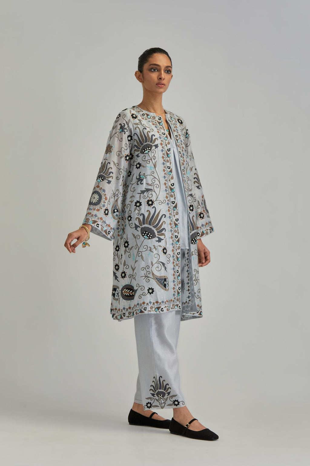 Blue short front open A-line jacket, fully embroidered with bold appliqué flowers, earthy-coloured Aari thread work and silk tassels, paired with Blue silk Chanderi straight pants with earthy coloured appliqué at hem.
