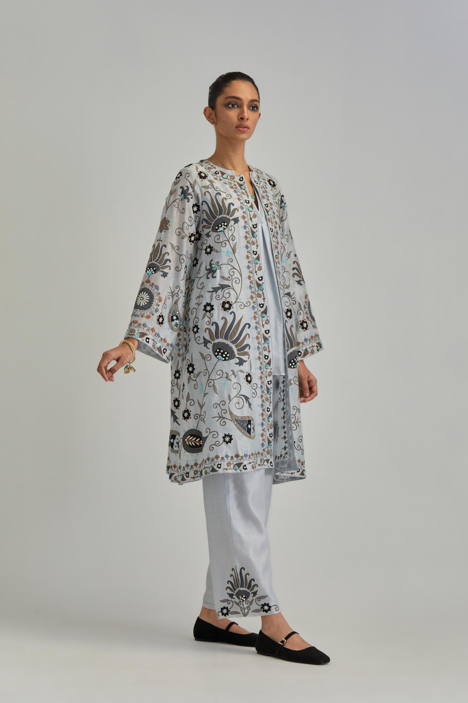 Blue short front open A-line jacket, fully embroidered with bold appliqué flowers, earthy-coloured Aari thread work and silk tassels, paired with Blue silk Chanderi straight pants with earthy coloured appliqué at hem.