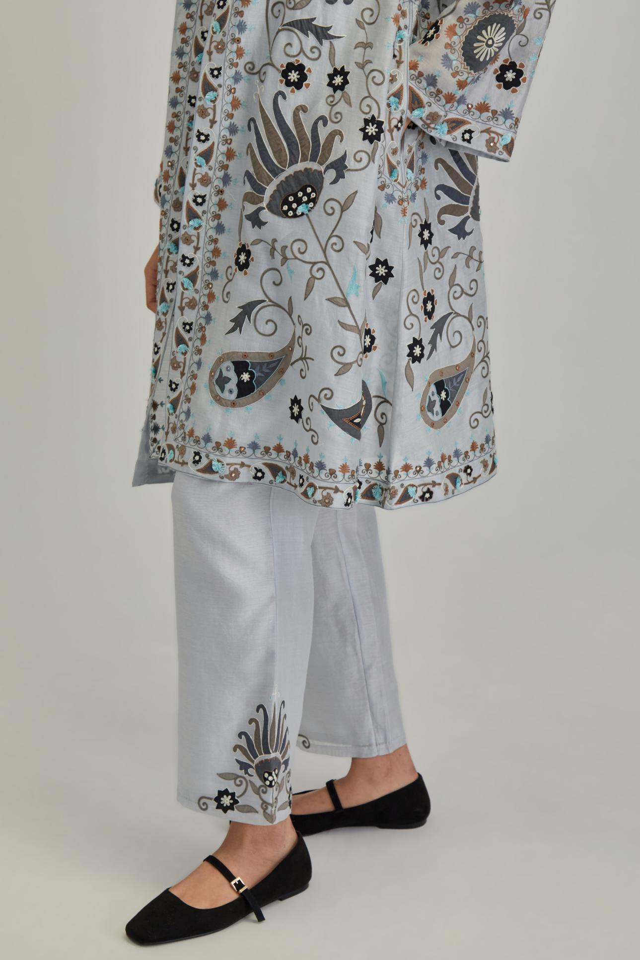 Blue short front open A-line jacket, fully embroidered with bold appliqué flowers, earthy-coloured Aari thread work and silk tassels, paired with Blue silk Chanderi straight pants with earthy coloured appliqué at hem.