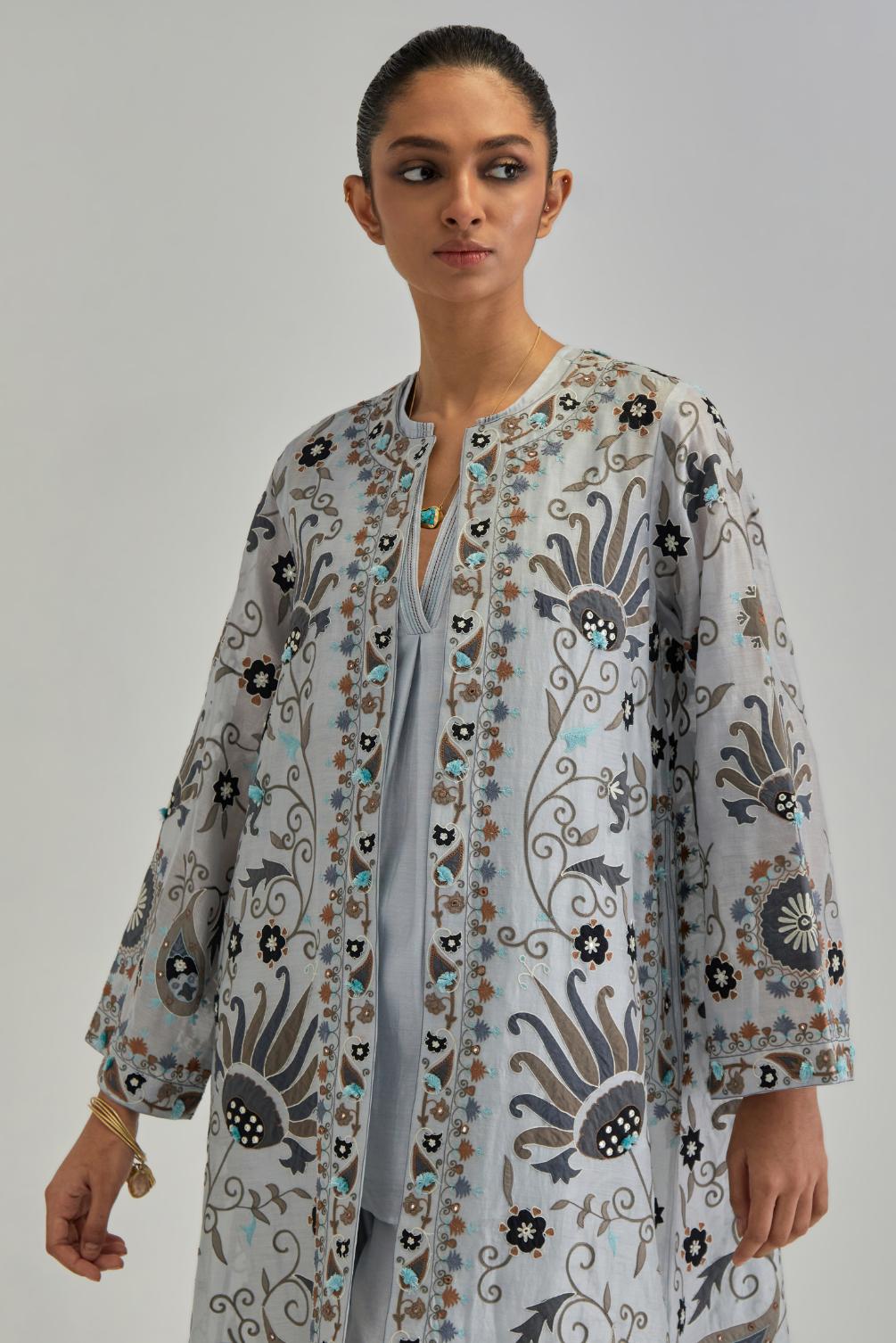 Blue short front open A-line jacket, fully embroidered with bold appliqué flowers, earthy-coloured Aari thread work and silk tassels, paired with Blue silk Chanderi straight pants with earthy coloured appliqué at hem.